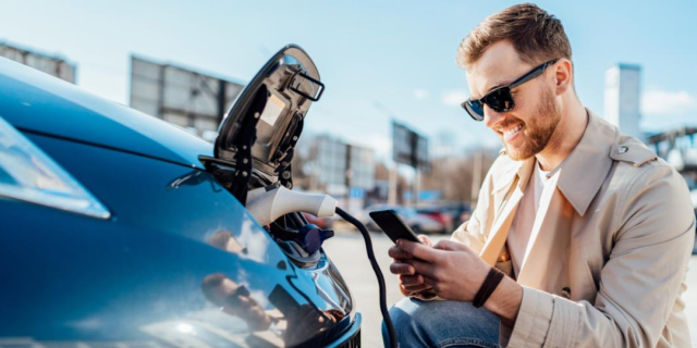 Mann mit Handy und Sonnenbrille beim Laden seines Elektroautos als Symbol für effizientes Fuhrparkmanagement