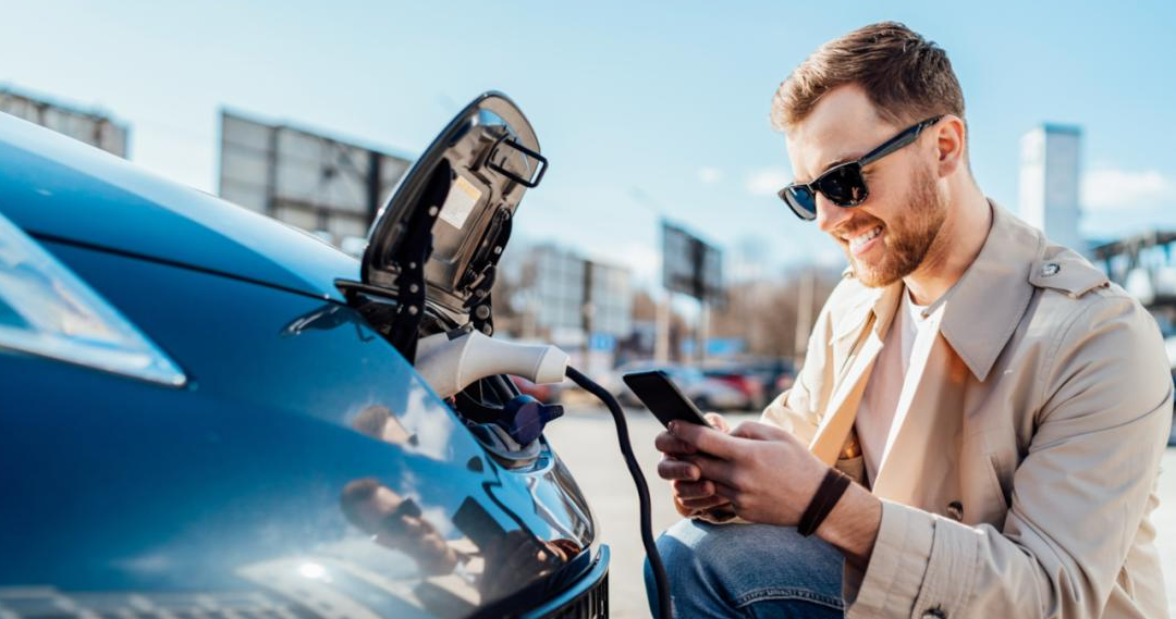 Mann mit Handy und Sonnenbrille beim Laden seines Elektroautos als Symbol für effizientes Fuhrparkmanagement